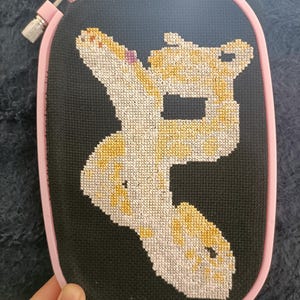Könnte beinhalten: Kreuzstich-Stickerei in einem rosa Stickrahmen. Das Design zeigt eine Giraffe in Gelb-, Weiß- und Beigetönen vor einem schwarzen Hintergrund. Die Giraffe befindet sich in einer spielerischen Pose. Der Rahmen wird von einer Hand gehalten.