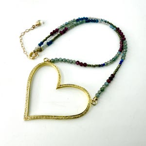 May include: A necklace featuring a large, open-frame gold heart pendant encrusted with small clear stones. The chain is composed of multi-colored faceted gemstone beads in shades of deep red, blue, green, and grey, finished with a gold clasp.