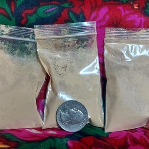 May include: Three small, clear plastic bags filled with a light tan powder. A silver coin rests in front of one of the bags. The bags are arranged on a red floral patterned fabric.