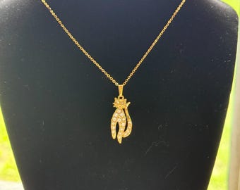May include: A gold-colored cat pendant necklace. The cat pendant is encrusted with small, clear crystals. The necklace is displayed on a black mannequin. The background is out of focus, with green and brown tones.