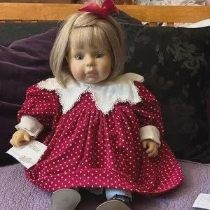 May include: A porcelain doll with blonde hair and a red bow. The doll wears a red polka-dot dress with a white collar and cuffs. The doll is holding a small white card.