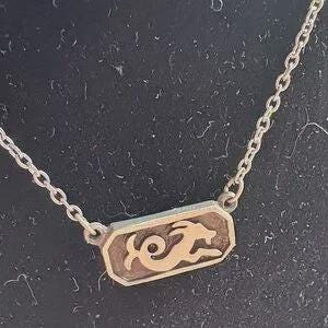 Könnte beinhalten: Eine silberfarbene Halskette mit einem rechteckigen Anhänger, der ein stilisiertes Bild einer springenden Ziege zeigt. Der Anhänger hat einen goldfarbenen Hintergrund und ist an einer filigranen Kette befestigt. Die Halskette wird vor dunklem Hintergrund gezeigt.