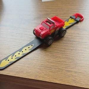 Op de afbeelding: Een rode speelgoedtruck-horloge met een zwarte band met een geel vlammenontwerp. Het horloge heeft een rode truck bovenop en een gespsluiting. Het horloge ligt op een houten oppervlak.
