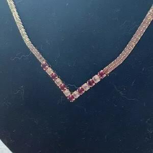 May include: A gold-toned necklace with a V-shaped pendant. The pendant is adorned with alternating red and clear gemstones. The necklace is displayed against a dark background.