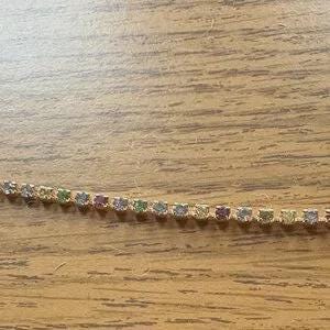 May include: A delicate gold-toned chain adorned with small, square-cut gemstones in a rainbow of colors. The gemstones are set closely together, creating a continuous line of sparkle. The chain is displayed on a light brown wooden surface.