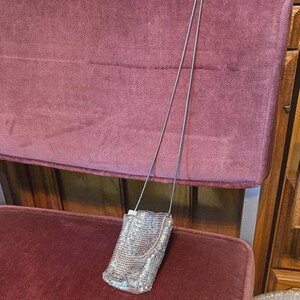 Puede incluir: Un pequeño bolso bandolera de malla plateada con una correa de cadena plateada larga y fina. El bolso tiene forma rectangular con cierre de solapa. El bolso cuelga contra una silla de terciopelo burdeos.