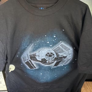 May include: Black t-shirt featuring a detailed graphic of a TIE fighter spacecraft against a starry, blue-toned space background. The shirt has a crew neck and short sleeves. The design is a tribute to a science fiction theme.