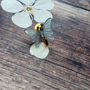 May include: A butterfly-shaped pin with translucent blue wings and a gold-colored body. The pin is set against a wooden background, with a white flower and a white petal. The pin is a decorative accessory.