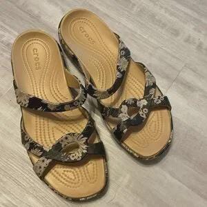 Könnte beinhalten: Ein Paar Crocs-Sandalen mit einem Blumenmuster in Schwarz, Grau und Beige. Die Sandalen haben ein beigefarbenes Fußbett und zwei Riemen über dem Fuß. Das Crocs-Logo ist sichtbar.