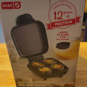 Op de afbeelding: Een zwarte Dash Deluxe Egg Bite Maker met vier kookbekers. De doos toont het apparaat met gekookte eierhapjes, een siliconen beker en tekst met 12 gram eiwit per portie.