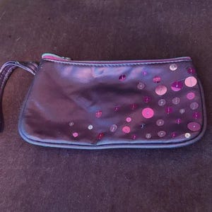 Puede incluir: Un pequeño bolso de mano morado oscuro con una correa de muñeca. El bolso rectangular está decorado con lentejuelas iridiscentes dispersas en tonos de rosa y morado. La cremallera es de un verde claro contrastante.