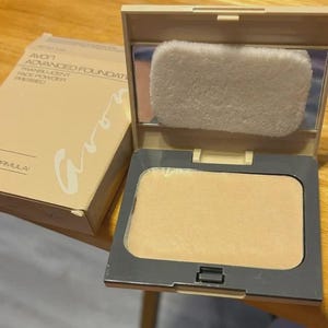 May include: Open beige compact of Avon Advanced Foundation Translucent Face Powder. The compact has a mirror and a powder puff. The pressed powder is a light beige colour. The box is beige with the Avon logo.