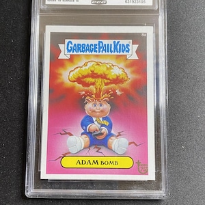 May include: A Garbage Pail Kids trading card, graded and encased. The card features an illustration of a cartoon character with a mushroom cloud explosion above its head. The card is labeled "ADAM BOMB".