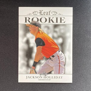 May include: A baseball card featuring Jackson Holliday, a shortstop, in an orange and black uniform. The card is labelled "Leaf Rookie" at the top. The card has a white border and a blurred background.