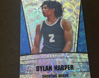 May include: A basketball trading card featuring Dylan Harper, a shooting guard, in a blue and white jersey with the number 7. The card has a holographic design with the word "ROOKIE" at the top.