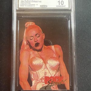 May include: A graded trading card featuring Madonna from the 1991 Rock Street #2 Prototype series. The card is encased in a protective holder and graded as Gem Mint 10 by CCG. The image on the card shows Madonna in a stage costume.