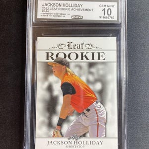 May include: A baseball card in a protective case. The card shows Jackson Holliday in an orange and black uniform. The card is labelled "2022 LEAF ROOKIE ACHIEVEMENT" and graded "GEM MINT 10". The card is a "ROOKIE" card.