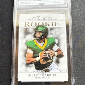 May include: A Dillon Gabriel rookie card from the 2024 Leaf Special Release, graded Gem Mint 10. The card features a photo of the quarterback in a green and yellow uniform, holding a football. The card is encased in a protective holder.