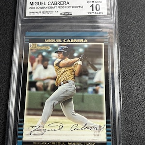 Puede incluir: Una tarjeta de béisbol de Miguel Cabrera en pose de bateo, protegida en un estuche. La tarjeta está calificada como GEM MINT 10. El texto incluye "2002 Bowman Draft Prospect" y "SE FLORIDA MARLINS".