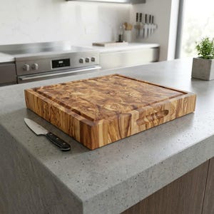 Puede incluir: Una tabla de cortar cuadrada y gruesa de madera de olivo con un patrón de veta natural prominente y asas laterales integradas, acompañada de un pequeño cuchillo de cocina con mango negro y hoja plateada, sobre una encimera de piedra gris.
