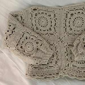 Könnte beinhalten: Ein beigefarbener, handgehäkelter Pullover mit rundem Ausschnitt und langen Ärmeln. Der Pullover hat ein sich wiederholendes kreisförmiges Muster am ganzen Körper und an den Ärmeln, mit einem gewellten Rand am unteren Saum. Das Kleidungsstück ist aus weichem, strukturiertem Garn gefertigt.
