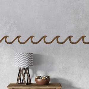 Puede incluir: Decoración de pared en forma de ola de madera color nogal. Debajo, una mesa de madera sostiene una lámpara con una pantalla con estampado de chevron gris y blanco, y un cuenco marrón con objetos decorativos. La pared es gris claro.