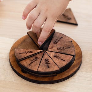 May include: A wooden fraction puzzle with a circular base and triangular pieces. Each piece is marked with the fraction 1/8. A hand is shown placing a piece into the puzzle. The puzzle is made of dark brown wood.