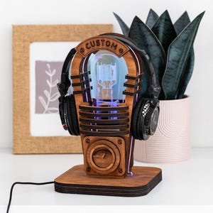Könnte beinhalten: Eine Holzlampe im Jukebox-Stil mit dem Wort "CUSTOM" oben. Die Lampe hat einen beleuchteten Innenraum und wird von schwarzen Kopfhörern flankiert. Der Sockel ist quadratisch und die Lampe steht auf einer weißen Oberfläche.