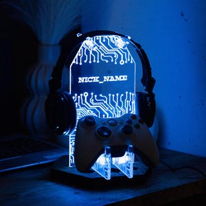 May include: A blue-lit gaming headset stand featuring a circuit board design and the text "NICK_NAME". A white game controller rests on the stand, which is made of wood. The stand is illuminated with blue light.
