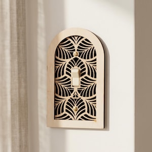 Könnte beinhalten: Eine dekorative Lichtschalterabdeckung mit gewölbtem Oberteil. Die Abdeckung besteht aus hellem Holz mit einem lasergeschnittenen floralen Design auf schwarzem Hintergrund. Ein cremefarbener Schalter ist zentriert. Goldfarbene Akzente sind sichtbar.