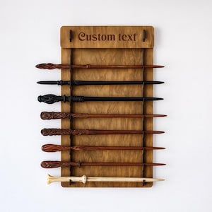 May include: Wooden wand display with seven wands of varying designs and colors. The wands are held in place by metal rods against a wooden backing. The top of the display has the words "Custom text" engraved.