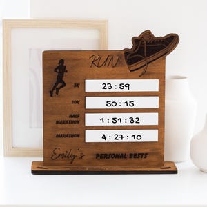 May include: Wooden running personal bests display board with a silhouette of a runner and a wooden shoe. The board has the word "RUN" and the text "Emily's PERSONAL BESTS". It displays times for 5K, 10K, half marathon, and marathon.