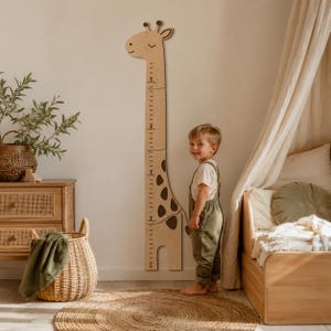 May include: A light-coloured wooden giraffe-shaped growth chart mounted on a wall. The chart features a printed scale with numbers 1 through 5, dark brown spots, and a smiling giraffe head at the top. The wood has a natural grain and smooth finish.