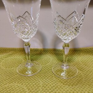 May include: Two clear crystal wine glasses with diamond-cut patterns on the upper portion of the bowl. The glasses have a classic design with a wide bowl, a slender stem, and a circular base. They are displayed on a textured, light green surface.