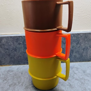 May include: A stack of three plastic mugs with square handles. The top mug is dark brown, the middle mug is bright orange, and the bottom mug is olive green. Each mug features a cylindrical body with a slightly flared rim and a matching handle.