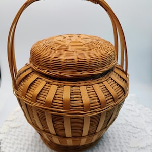 May include: A round, woven basket with a lid and a handle. The basket is made of light brown bamboo or wicker, with a tightly woven pattern on the body and lid. The handle is arched and attached to the sides. The basket sits on a white lace surface.