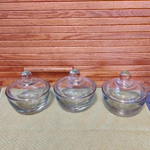 May include: Three clear glass bowls with matching lids. Each bowl has a rounded shape and a small, round knob on top of the lid. The bowls are sitting on a light green surface, with a wooden background.