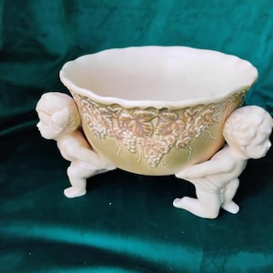 May include: Cream-colored ceramic bowl with a scalloped rim, supported by two cherubic figures. The bowl's exterior features a relief design of grapevines and leaves. The figures are kneeling and appear to be holding up the bowl. The background is a dark green.