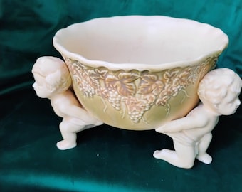 May include: Cream-colored ceramic bowl with a scalloped rim, supported by two cherubic figures. The bowl's exterior features a relief design of grapevines and leaves. The figures are kneeling and appear to be holding up the bowl. The background is a dark green.