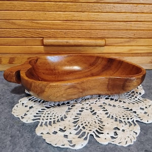 May include: A wooden, heart-shaped bowl with a handle on the left side. The bowl is made of a light brown wood and sits on a white, lacy doily. The background features a wooden breadbox.