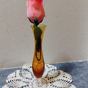 May include: A pink rose in a slender amber glass vase. The vase sits on a white, lacy doily. The rose has a gradient of pink hues, and the vase has a long, elegant shape. The background is a neutral color.