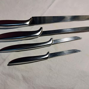 May include: A set of four stainless steel knives with curved, dark-colored handles. The blades vary in size, with one having a forked tip. The knives are arranged on a white surface, showcasing their polished finish.