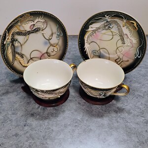 May include: A set of two teacups and saucers. The white cups have gold handles and ornate dragon designs. The saucers feature a black border with a dragon motif and a cream-colored center with pink accents. The cups sit on small, round, dark brown bases.