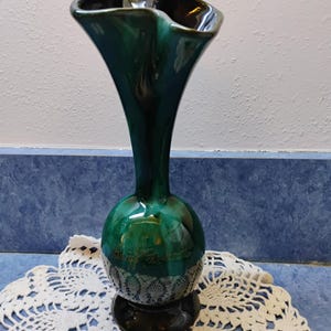 May include: A tall, green ceramic vase with a flared, ruffled top and a bulbous midsection. The vase has a dark base and a decorative lace pattern near the base. The vase has the text "Daryl Prescott" on the side.