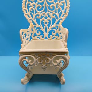 May include: Ivory decorative rocking chair with a heart-shaped back and ornate scrollwork. The chair has a small rectangular seat and curved rockers. The background is a solid blue.