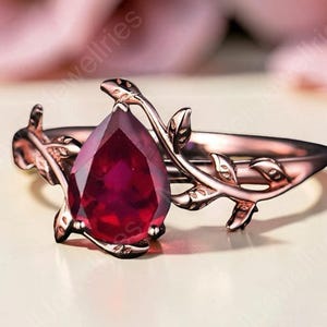 May include: A rose gold ring featuring a pear-shaped deep red gemstone as the centre stone. The band is designed with a detailed vine and leaf motif that wraps around the finger, extending from the base of the central red teardrop-shaped stone.