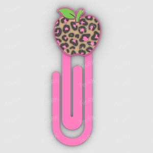 Puede incluir: Un clip de papel rosa con una parte superior en forma de manzana. La manzana tiene un diseño de estampado de leopardo en marrón, negro y rosa, con una hoja verde. El clip de papel es de color rosa vibrante.