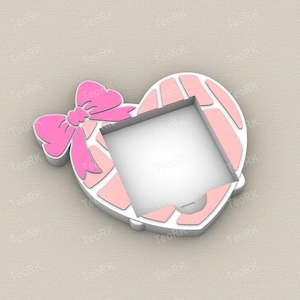 May include: A pink heart-shaped cookie cutter with a bow on the left side. The heart has a white border and a square cutout in the center. The bow and the heart's interior are shades of pink.