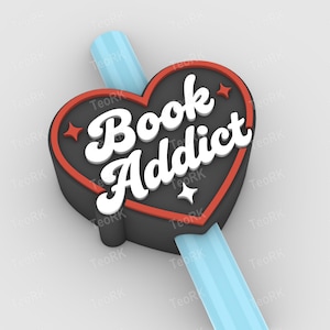 Puede incluir: Un marcapáginas en forma de corazón con la inscripción "Book Addict" en blanco. El marcapáginas tiene una base negra con un borde rojo y una cinta azul claro. Se ven pequeños detalles de estrellas.