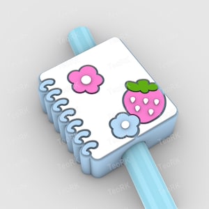 Op de afbeelding: Een lichtblauwe strooitopper in de vorm van een notitieboekje met een witte kaft versierd met een roze bloem, een aardbei en een blauwe bloem. De strooitopper is verbonden met een lichtblauw rietje.